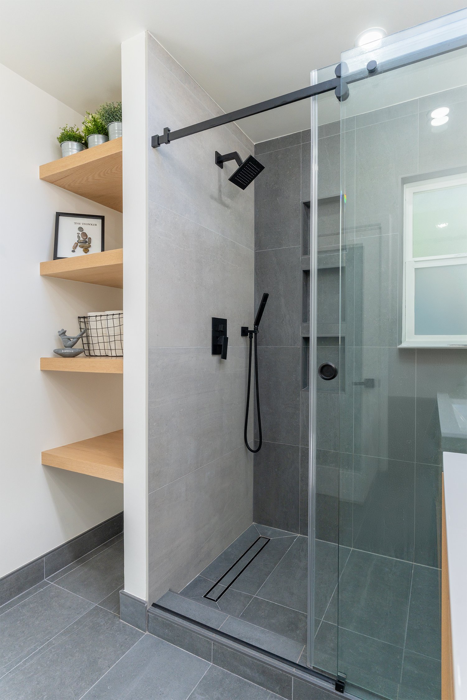 Modern bathroom remodel in Los Altos, CA, with a glass shower enclosure, gray tiled walls, black rainfall showerhead, and light wood floating shelves for a sleek and contemporary design