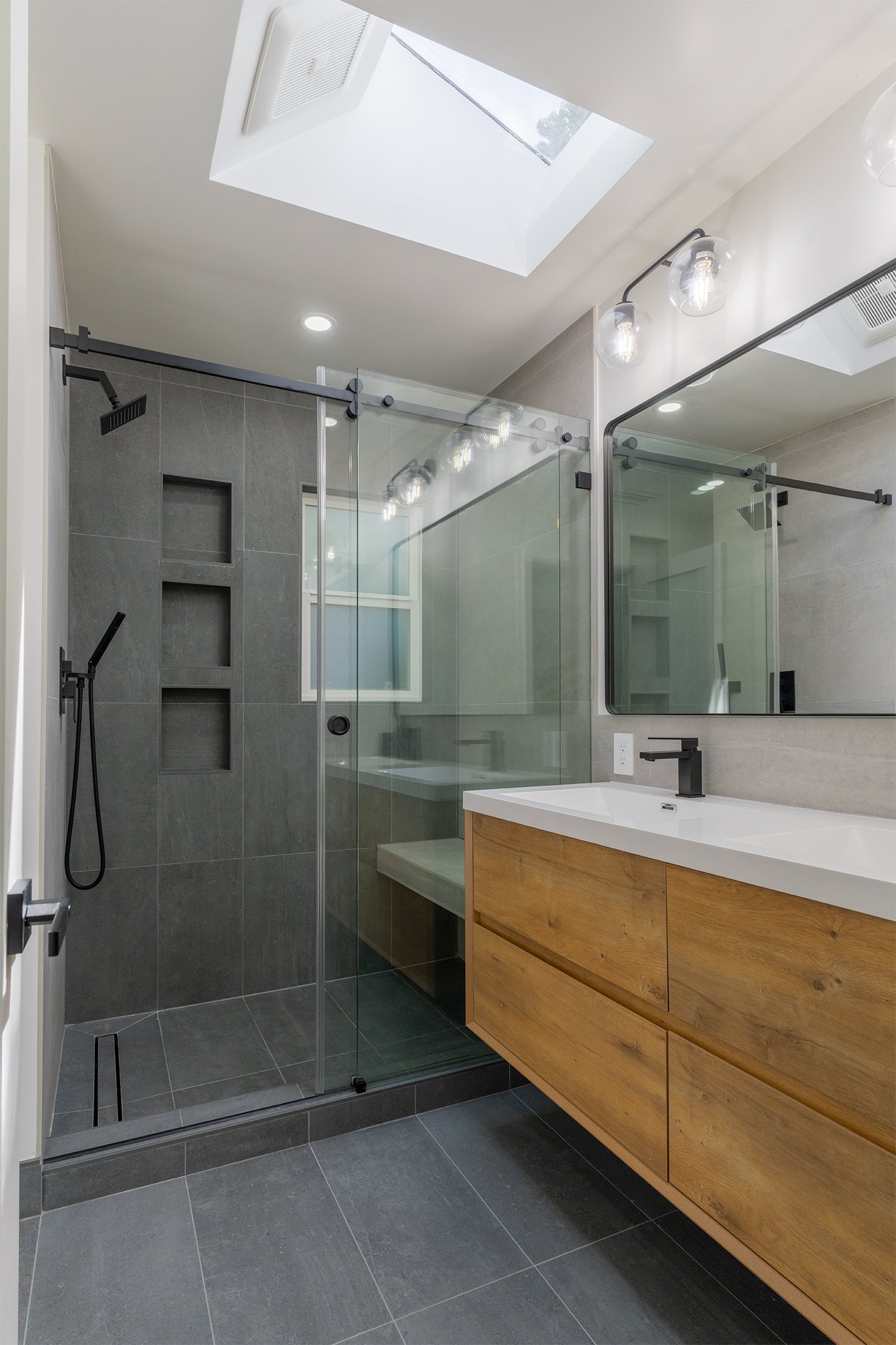 Contemporary bathroom remodel in Los Altos, CA, featuring a floating wood vanity, gray tiled shower with built-in niches, frameless glass enclosure, and natural light from a skylight.