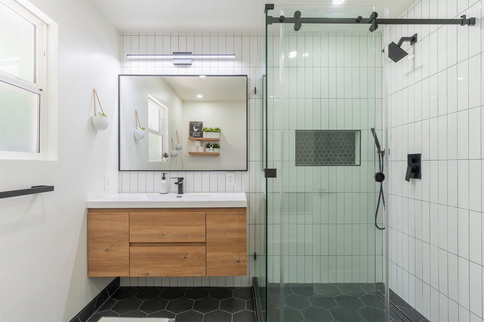 Modern bathroom remodel in Los Altos, CA, featuring a floating wood vanity, frameless glass shower with white vertical subway tiles, black hexagon tile floor, and sleek black fixtures.