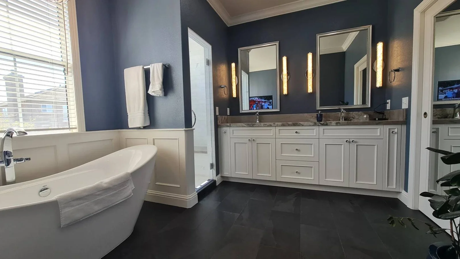 Luxury bathroom remodel in San Jose, CA, featuring a freestanding soaking tub, double vanity with white cabinets, elegant wall sconces, and dark tile flooring.